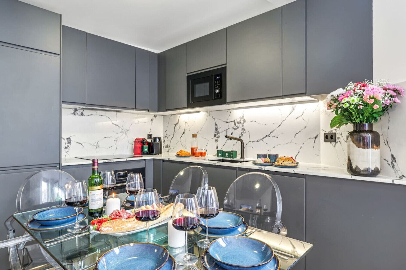 Een moderne keuken met grijze kastjes, een marmeren spatwand en een eethoek met blauwe borden en wijnglazen.