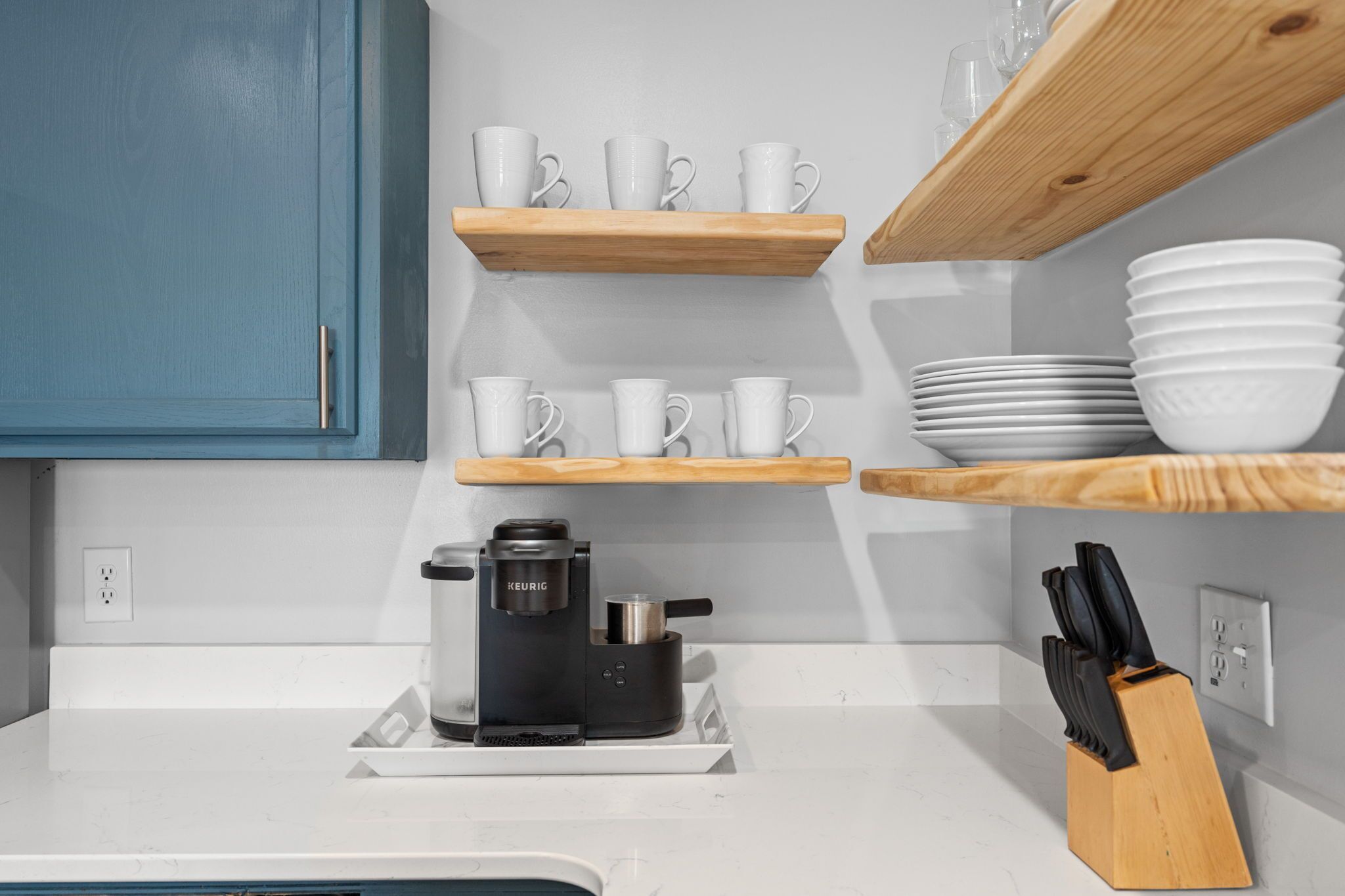 Une cuisine avec des plans de travail blancs, une machine à café et des étagères en bois où sont posées des tasses et des assiettes.