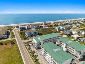 Vista aérea de una zona residencial costera con un edificio de varios pisos destacado, que tiene techo verde, rodeado de casas de una o dos plantas, una playa y un cielo azul despejado.