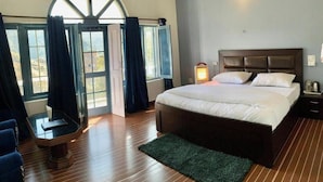 A bedroom with a large bed, a wooden floor, a green rug, a nightstand, a lamp, and a window with curtains.