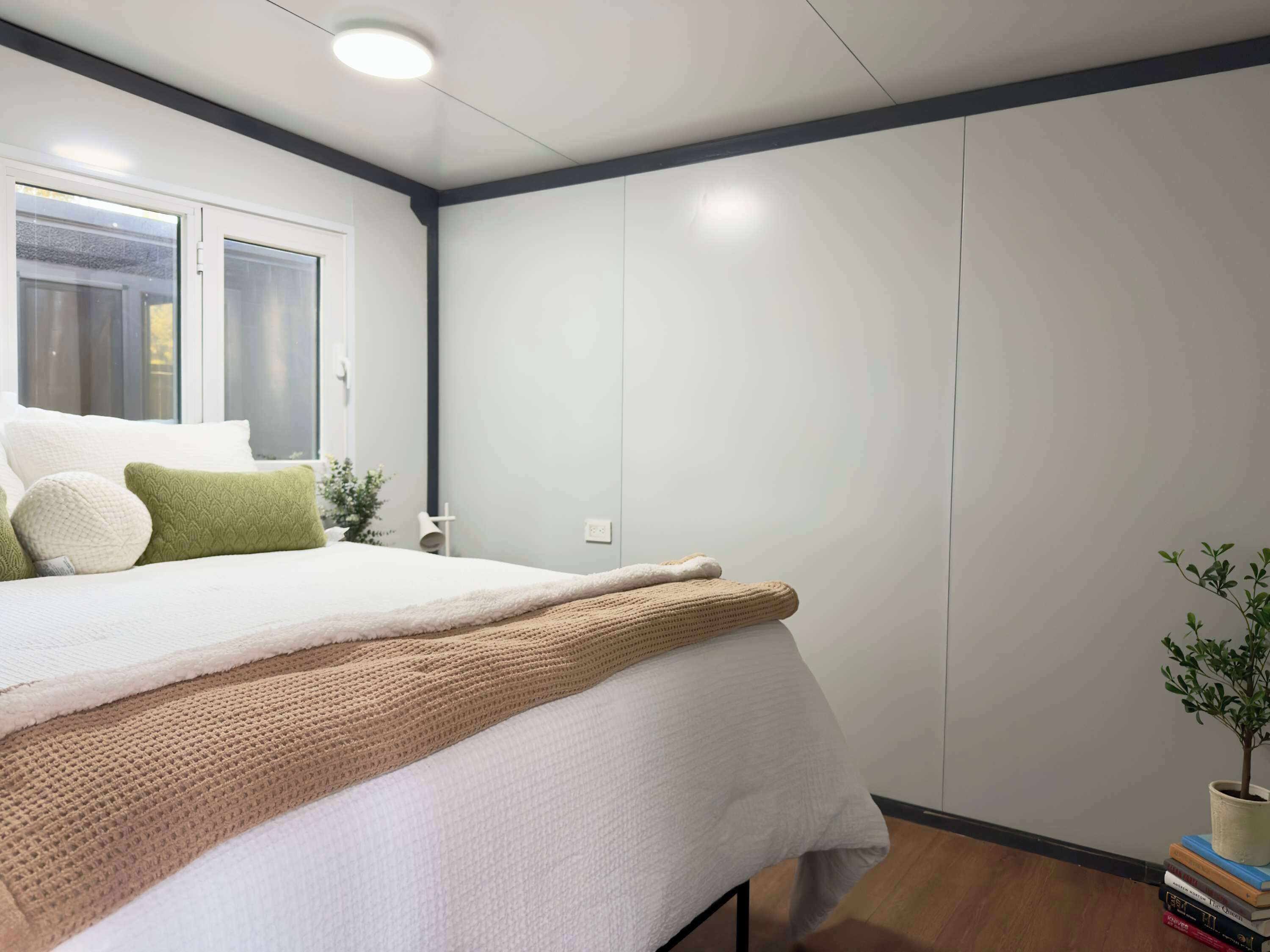 Ein Schlafzimmer mit einem Bett, Kissen, einer Decke, einer Topfpflanze und einem Bücherstapel.