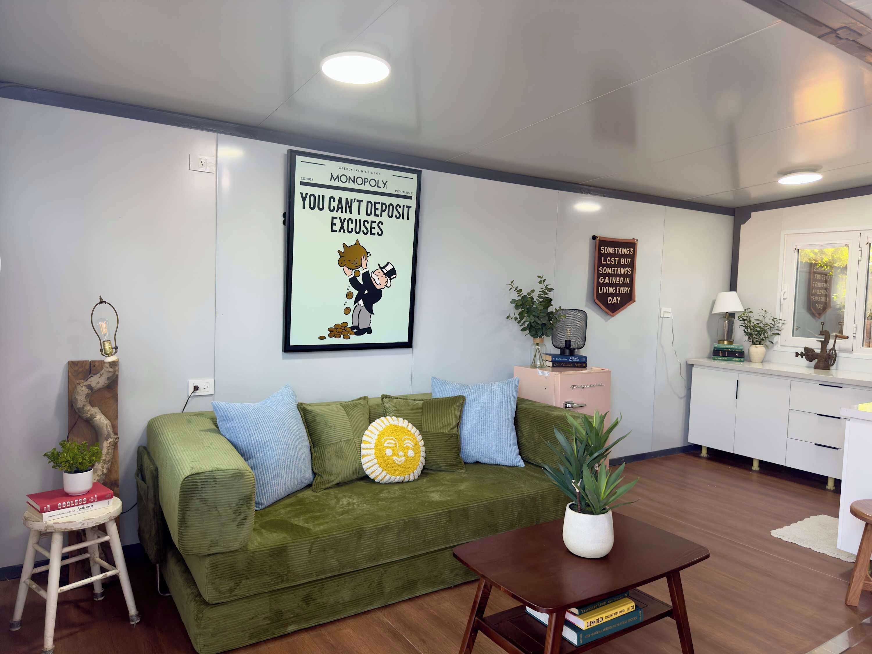 A living room with a green sofa, a coffee table, a framed poster, a small stool with a plant, and a window with a view.