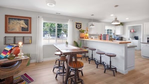 Una cocina con una isla central, taburetes de bar, iluminación colgante y un ventanal con cortinas.
