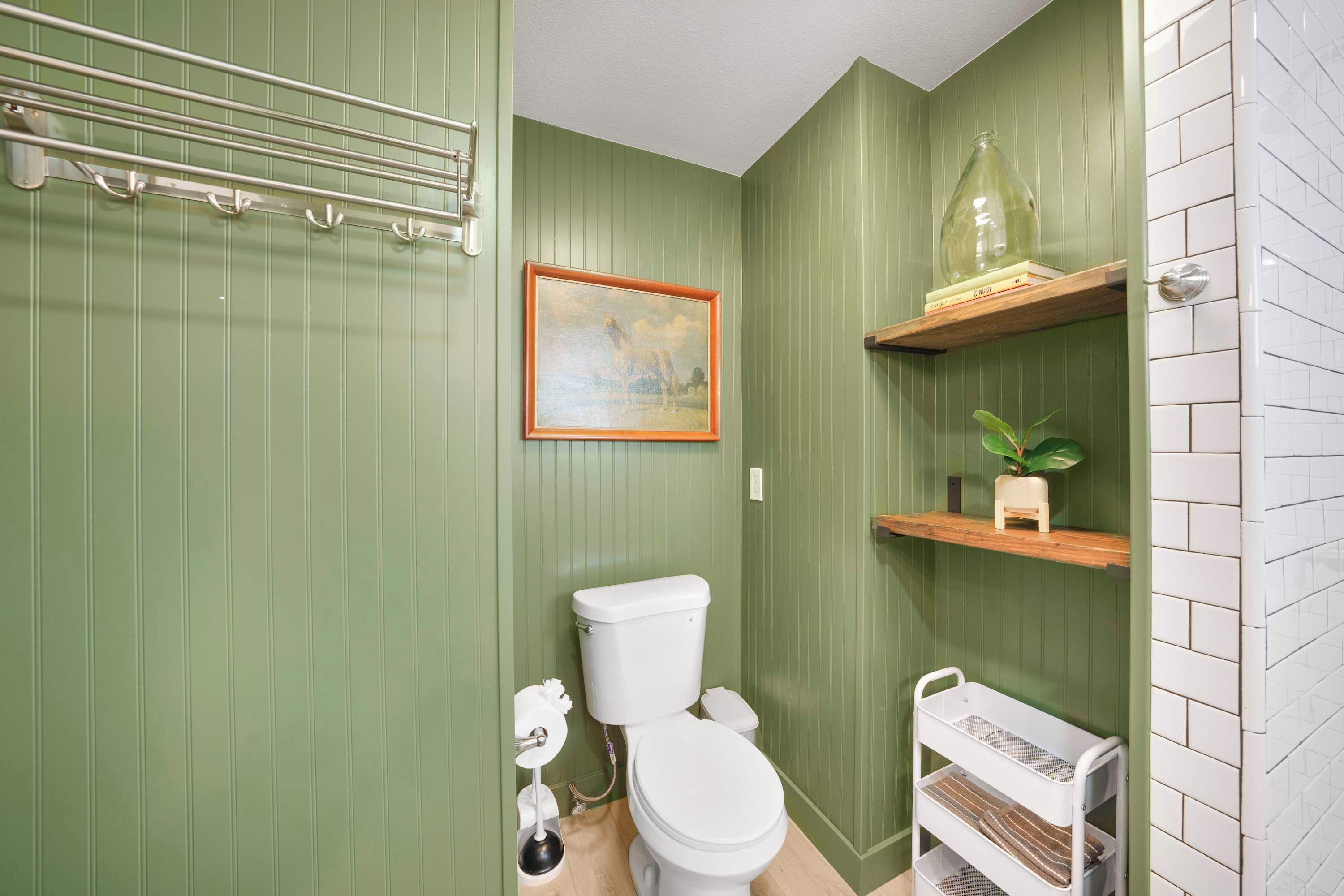 Ein Badezimmer mit grünen Wänden, einer weißen Toilette, einem gerahmten Bild und einem Regal mit einer Pflanze.