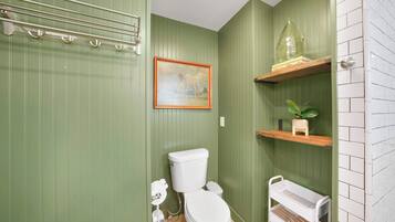 Ein Badezimmer mit grünen Wänden, einer weißen Toilette, einem gerahmten Bild und einem Regal mit einer Pflanze.