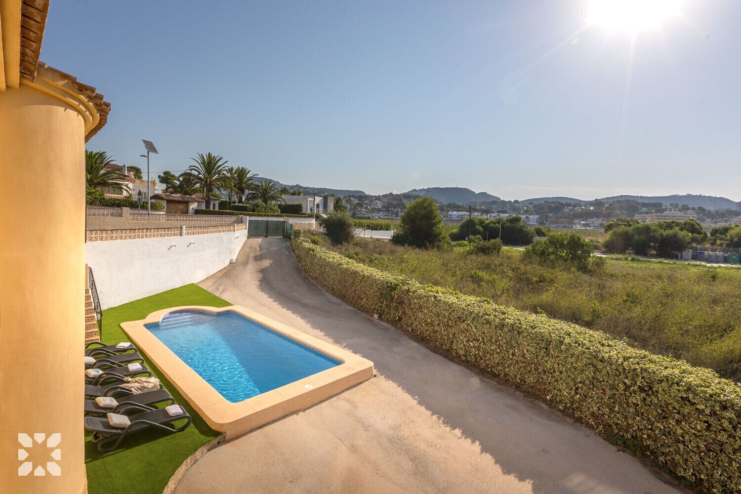 A swimming pool with a paved deck, a green lawn area, and a view of a residential area with houses and hills in the background.