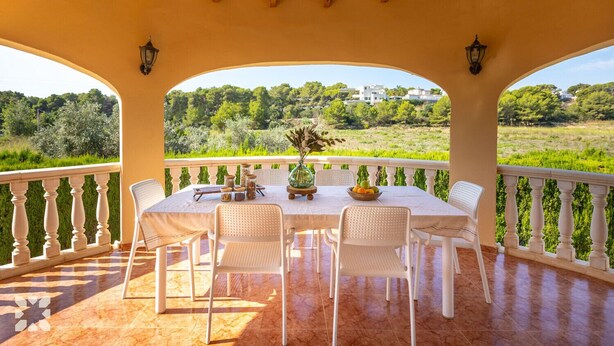 Un espacio exterior techado para comer con una mesa dispuesta para cuatro personas, rodeada de sillas blancas, con vistas a un paisaje verde y exuberante.