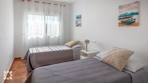 Un dormitorio con dos camas, una mesita de noche, una lámpara y una ventana con cortinas translúcidas.
