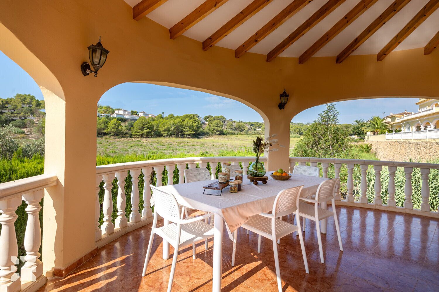 A covered outdoor patio with a table set for dining, surrounded by white chairs, and overlooking a lush landscape.