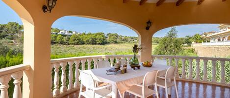 A covered outdoor patio with a table set for dining, surrounded by white chairs, and overlooking a lush landscape.