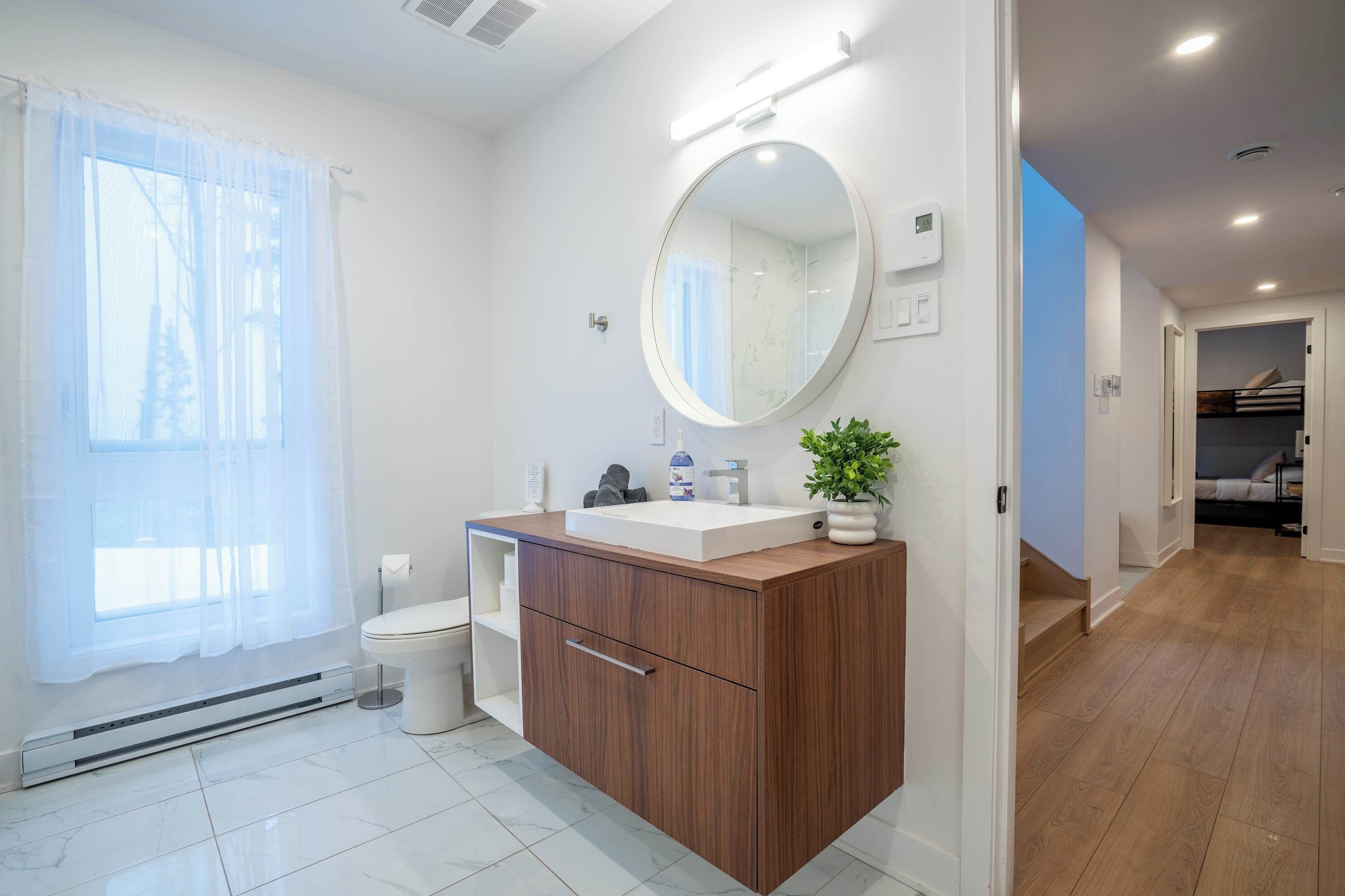 Een moderne badkamer met een wit toilet, een houten wastafelmeubel met een spiegel, en een kleine plant op de aanrecht.