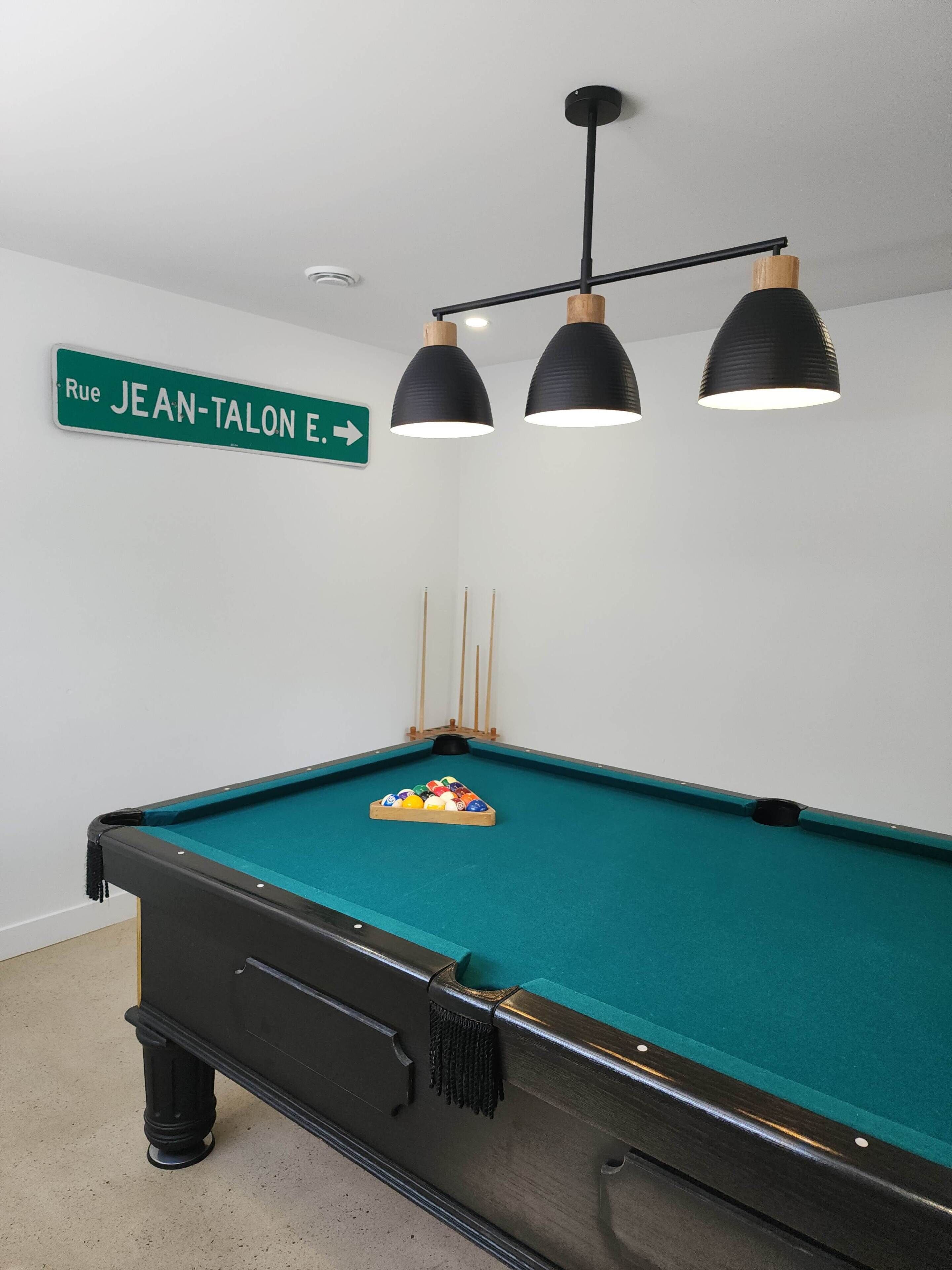 A pool table with a cue ball rack, overhead lighting, and a directional sign on the wall.
