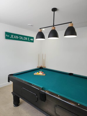 Une table de billard avec un porte-billes, un éclairage suspendu et un panneau indicateur sur le mur.