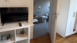 Un salón moderno con un televisor de pantalla plana sobre un mueble blanco, un control remoto y una revista en un estante. Un suelo de madera en espiga y una puerta blanca dan paso a un baño con lavamanos y bidé.