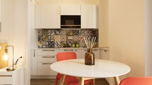 Una cocina compacta con armarios blancos, un salpicadero estampado, un microondas y una mesa redonda con sillas rojas.