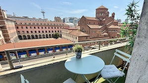 Un balcón con una mesa redonda y dos sillas blancas con cojines, con vistas a un paisaje urbano que incluye edificios históricos y una estructura moderna al fondo.
