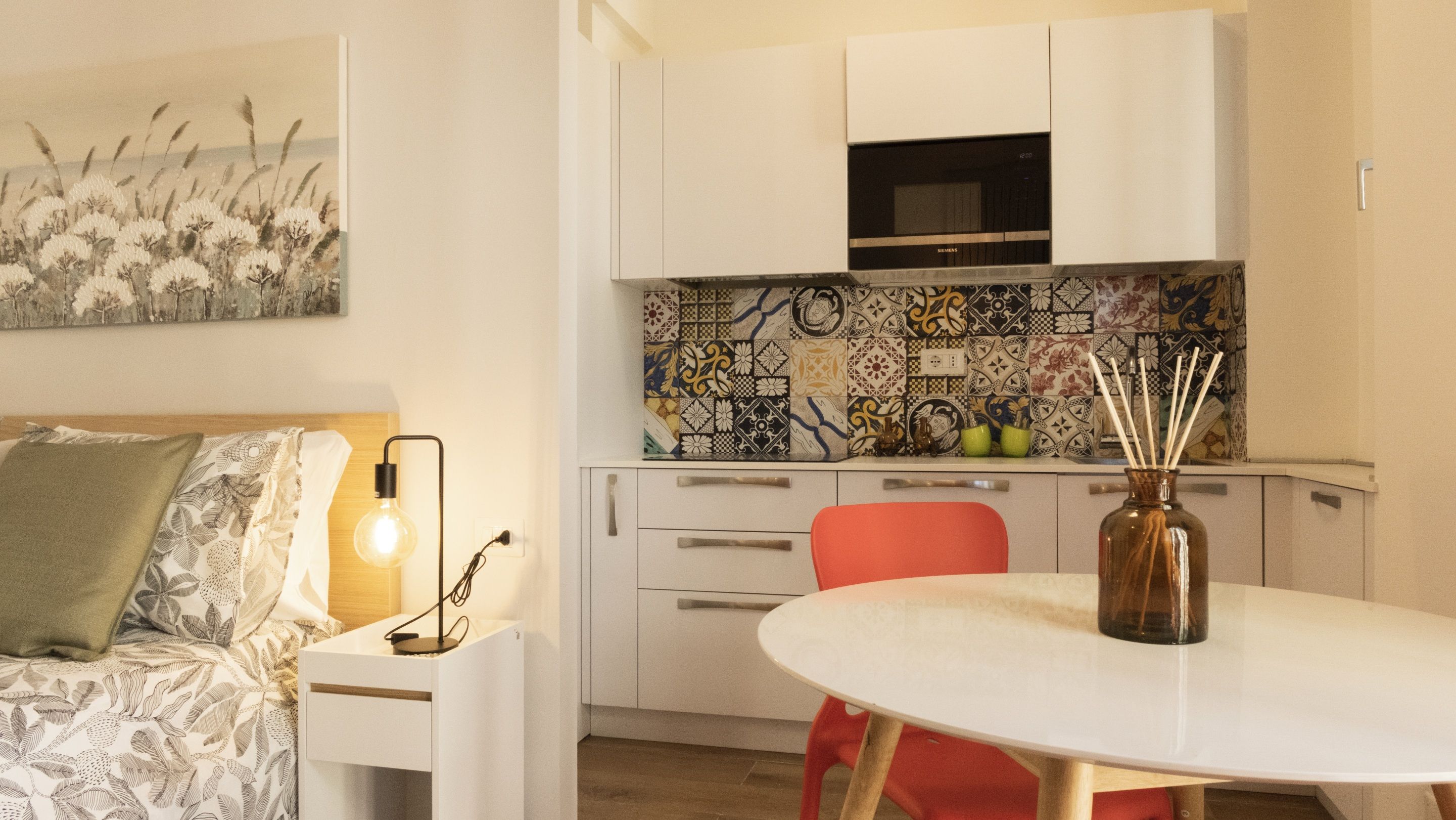 A compact kitchen with white cabinets, a microwave, and a colorful backsplash. There's a dining table with a vase and candles.