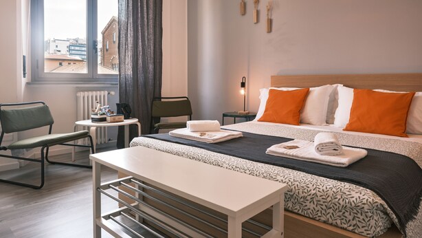 Una habitación de hotel moderna con una cama grande, una mesita de noche, una silla, una pequeña mesa y vistas a edificios al exterior.