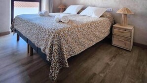 A bedroom with a wooden ceiling, a bed with a patterned bedspread, two bedside tables with lamps, and a large window with curtains. - Lodging fuerte trece (Encinasola)