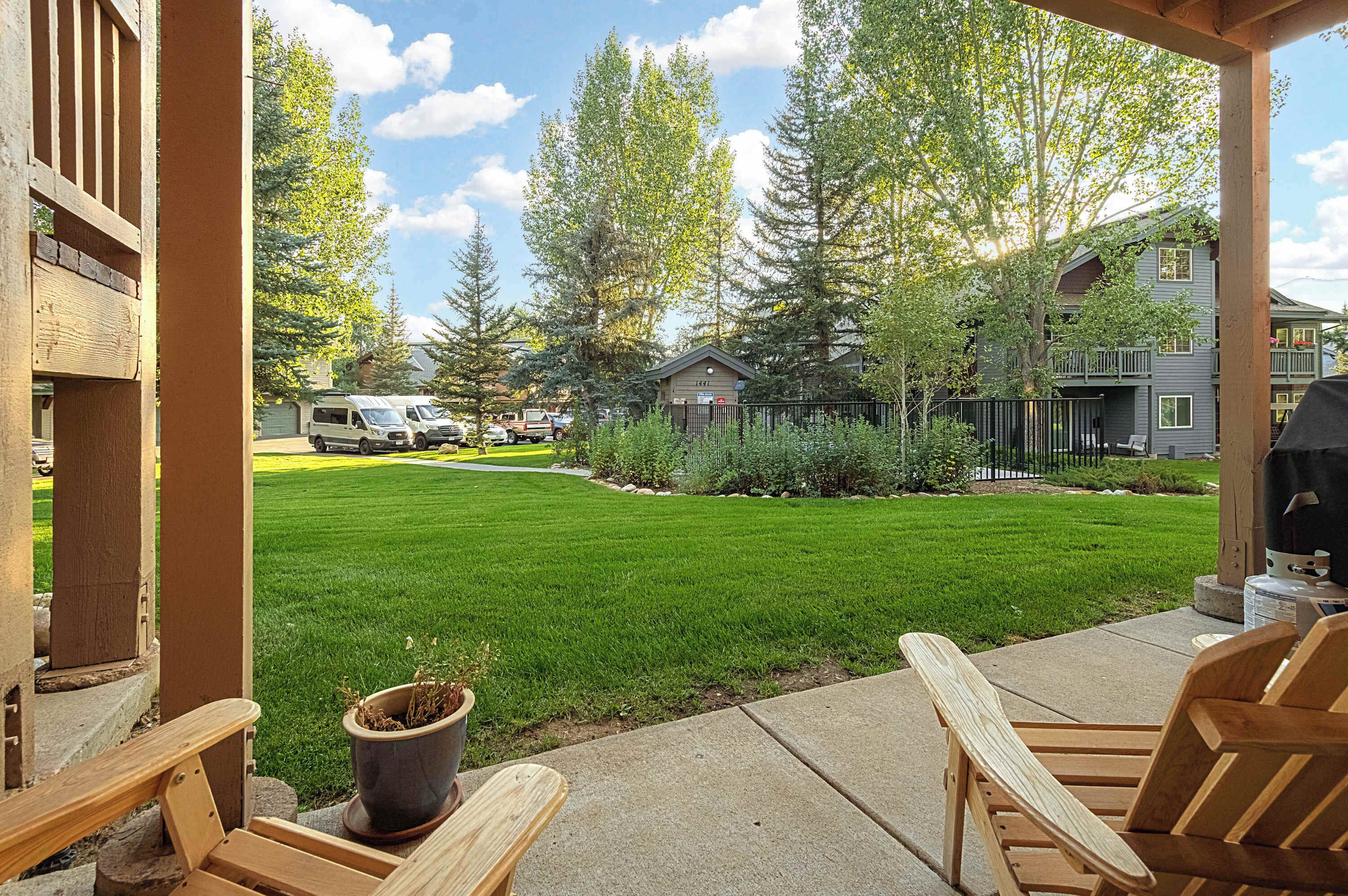 Une terrasse avec des chaises en bois et un barbecue, donnant sur une pelouse bien entretenue et des immeubles résidentiels.