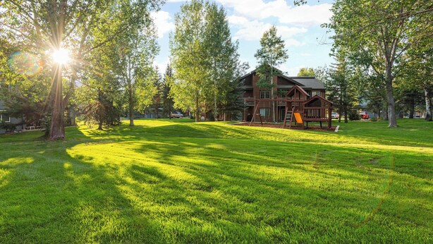 A large, rustic log cabin with a deck and a swing, surrounded by lush green grass and trees, under a clear sky.