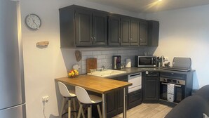 Cocina compacta con gabinetes oscuros, microondas, cafetera y un lavamanos. Hay dos sillas blancas en la encimera.
