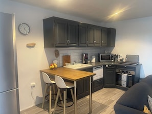 A compact kitchen with dark cabinets, a microwave, a coffee maker, and a sink. There are two white chairs at the kitchen counter.