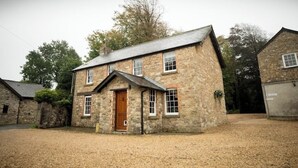 A stone building with a wooden door and windows, surrounded by a gravel driveway and trees. - The Coach House (Reynoldston)