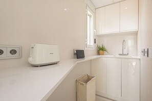 Une cuisine moderne avec des meubles blancs, un grille-pain encastré, un petit évier et une fenêtre qui laisse entrer la lumière naturelle.