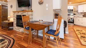 Un salón acogedor con chimenea, mesa de comedor de madera y sillas, un televisor montado en la pared, y una cocina visible a través de un vano abierto.