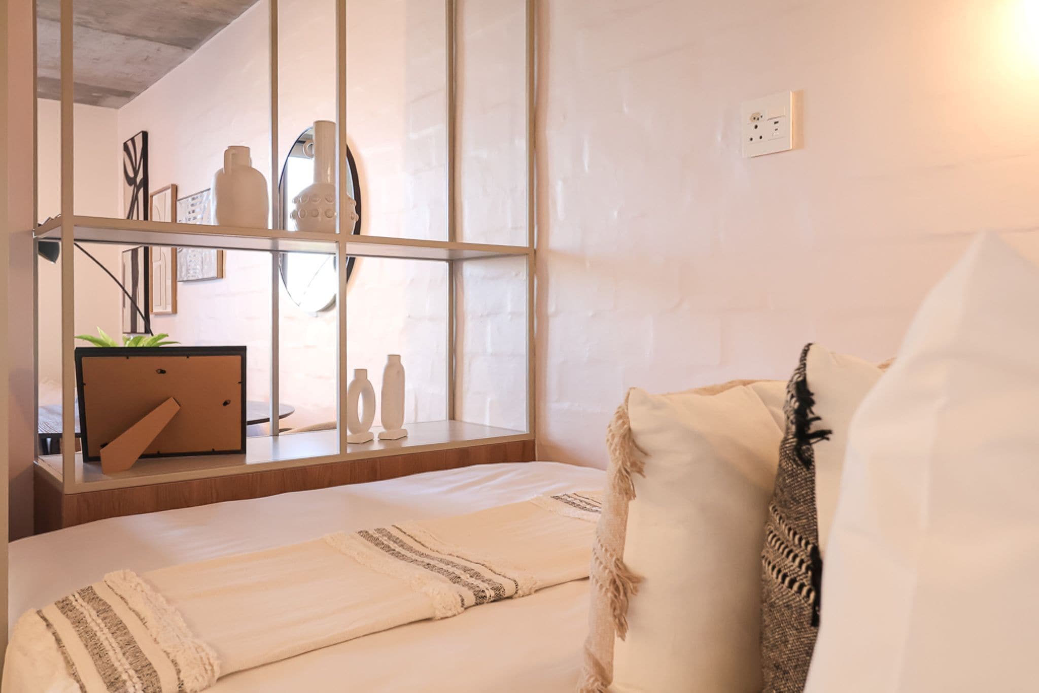 A bedroom with a bed, a bedside table with a plant, a mirror, and a shelf with decorative items.