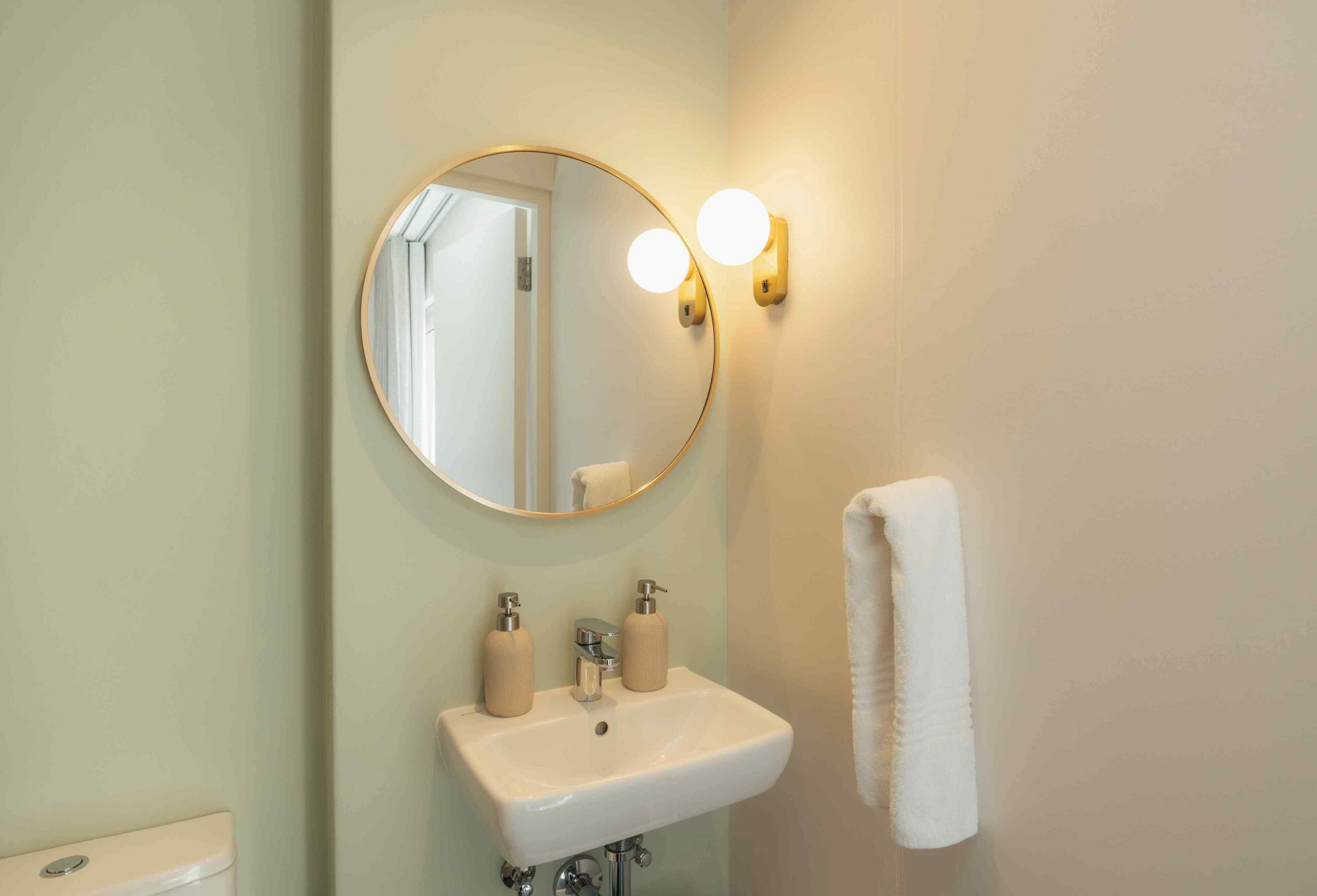 A bathroom with a round mirror, two wall-mounted lights, a sink with soap dispensers, and a white towel hanging on the wall.