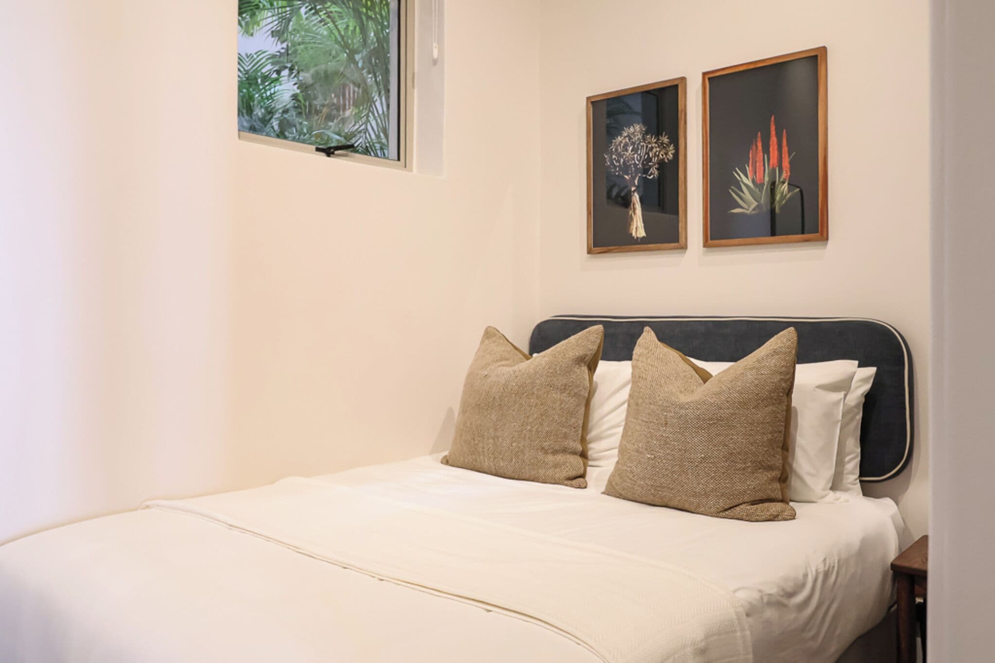 A neatly made bed with a dark headboard, two framed floral artworks on the wall, and a window with a view of greenery.
