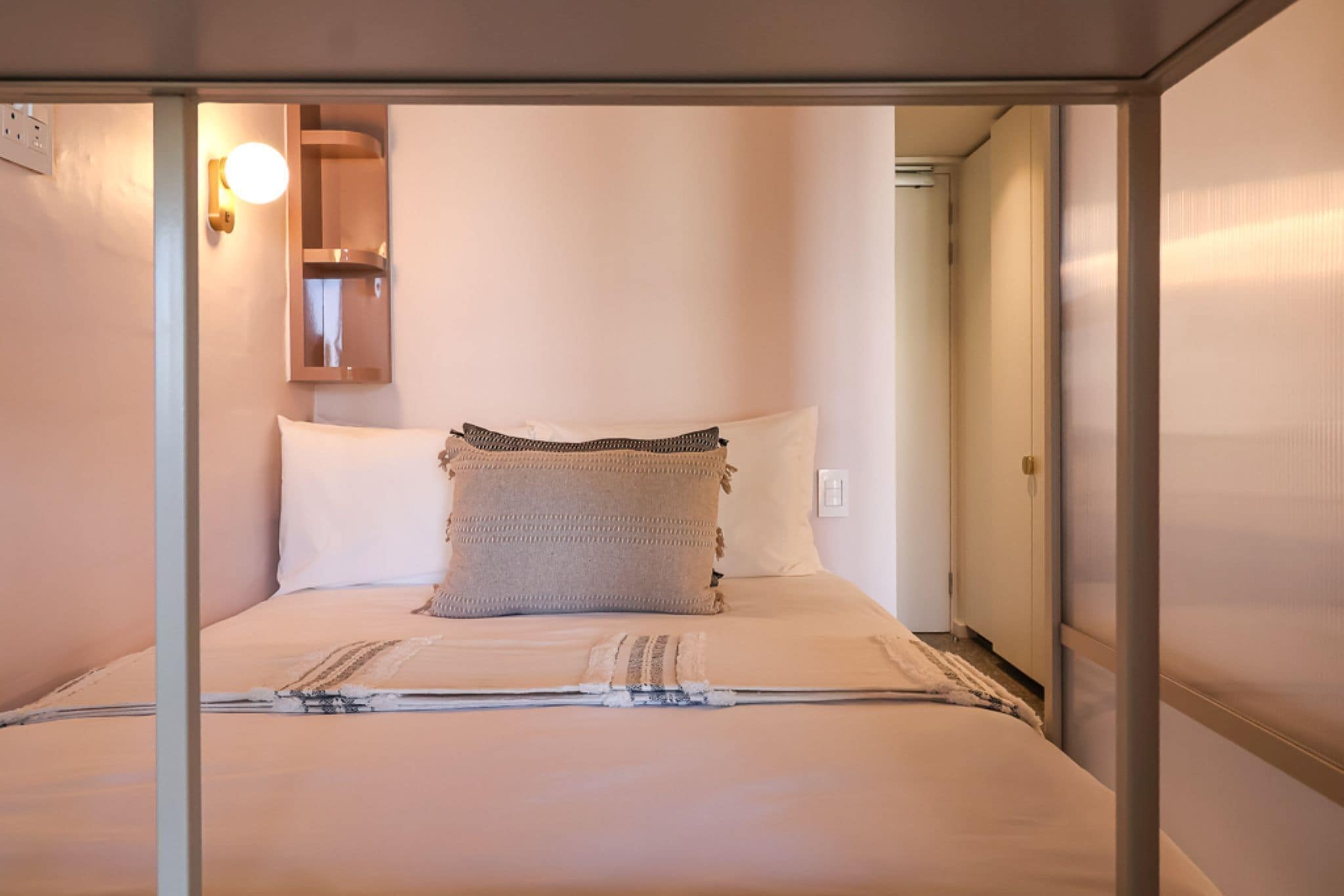 A neatly made bed with white and beige bedding, a wall-mounted light fixture, and a door leading to another room.