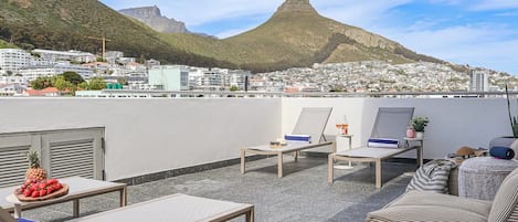 A rooftop terrace with lounge chairs, a table with fruit, and a view of a cityscape and mountain.