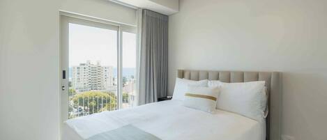 A modern hotel room with a large bed, a view of the city through a sliding door, and a neutral color scheme.
