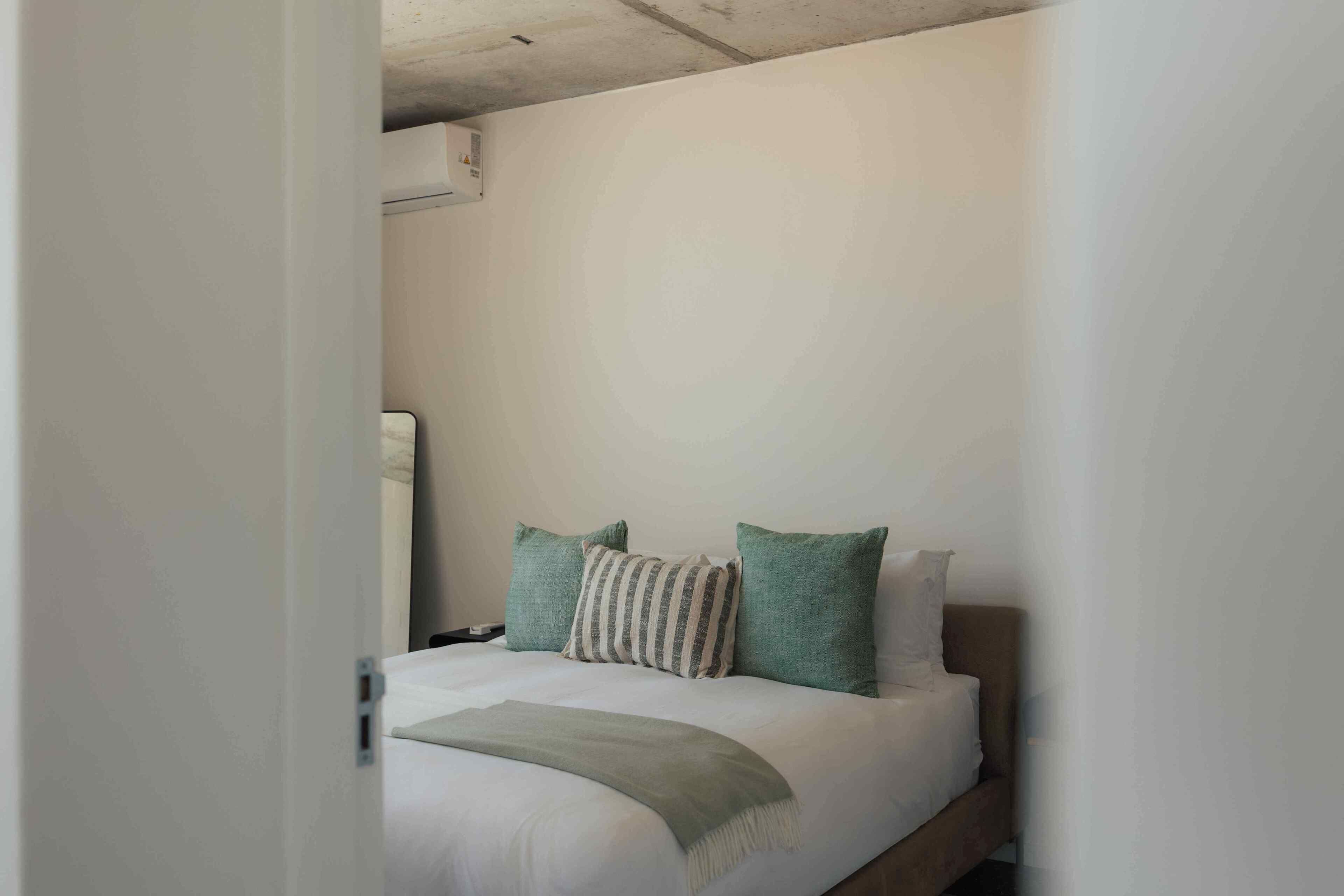 Cama bem arrumada com edredom branco e duas almofadas, uma listrada e outra verde lisa, em quarto simples com paredes brancas e cabeceira de madeira.