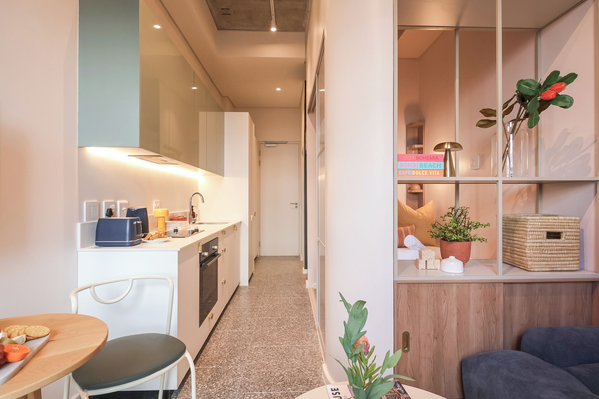 A compact kitchen with a dining area, a small table with a chair, and a sofa. There are cabinets, a microwave, and a coffee machine.