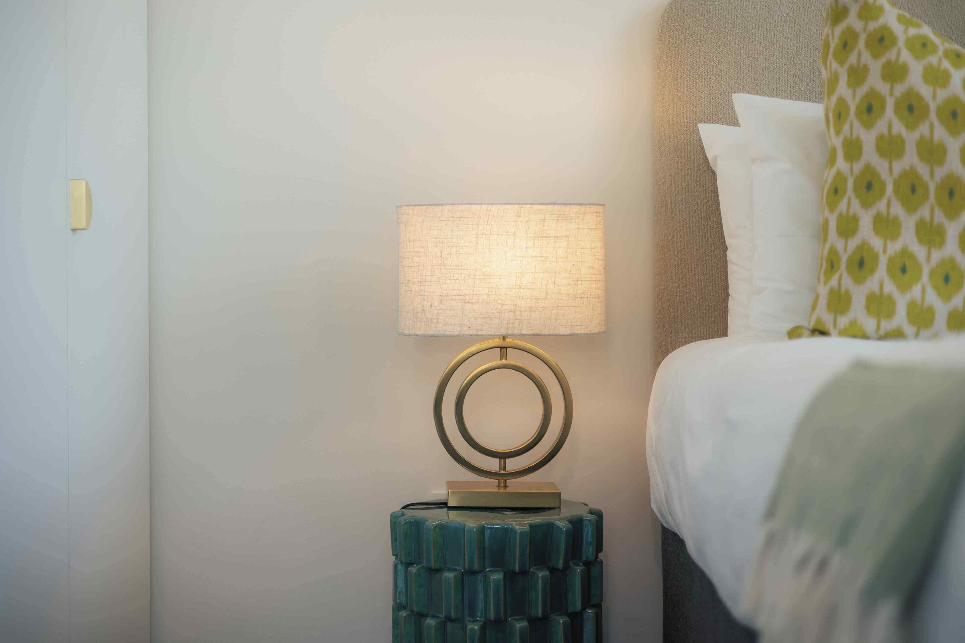 A bed with white bedding and a green patterned pillow, a wall-mounted lamp with a circular base, and a textured blue side table.