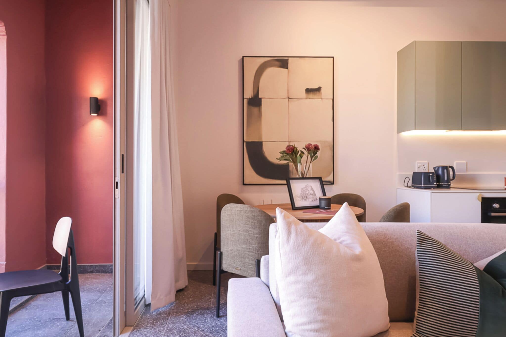 A modern hotel room with a sofa, a dining table with chairs, a large mirror, and a kitchenette.