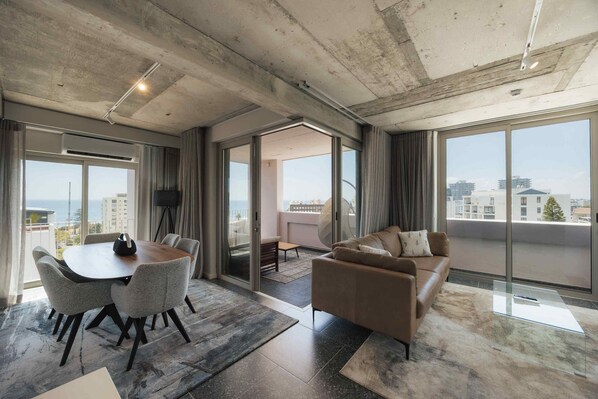 A modern living room with a large window, a sofa, a coffee table, and a dining area with chairs and a table. - ONEHUNDRED on M - Fluent Serviced Apartments (Cape Town)
