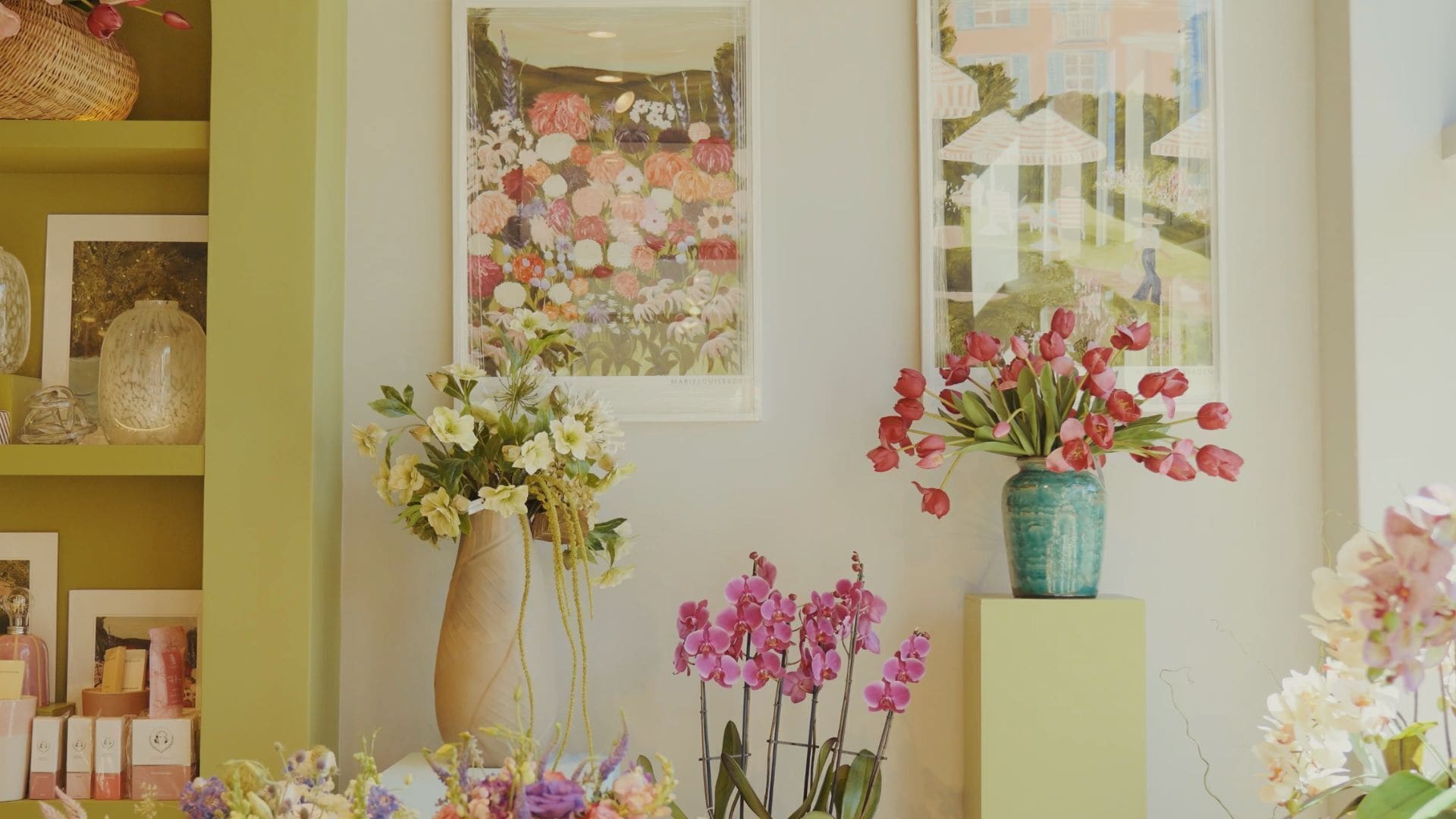 A floral display with various flowers in vases, a framed artwork, and a window view of a house.