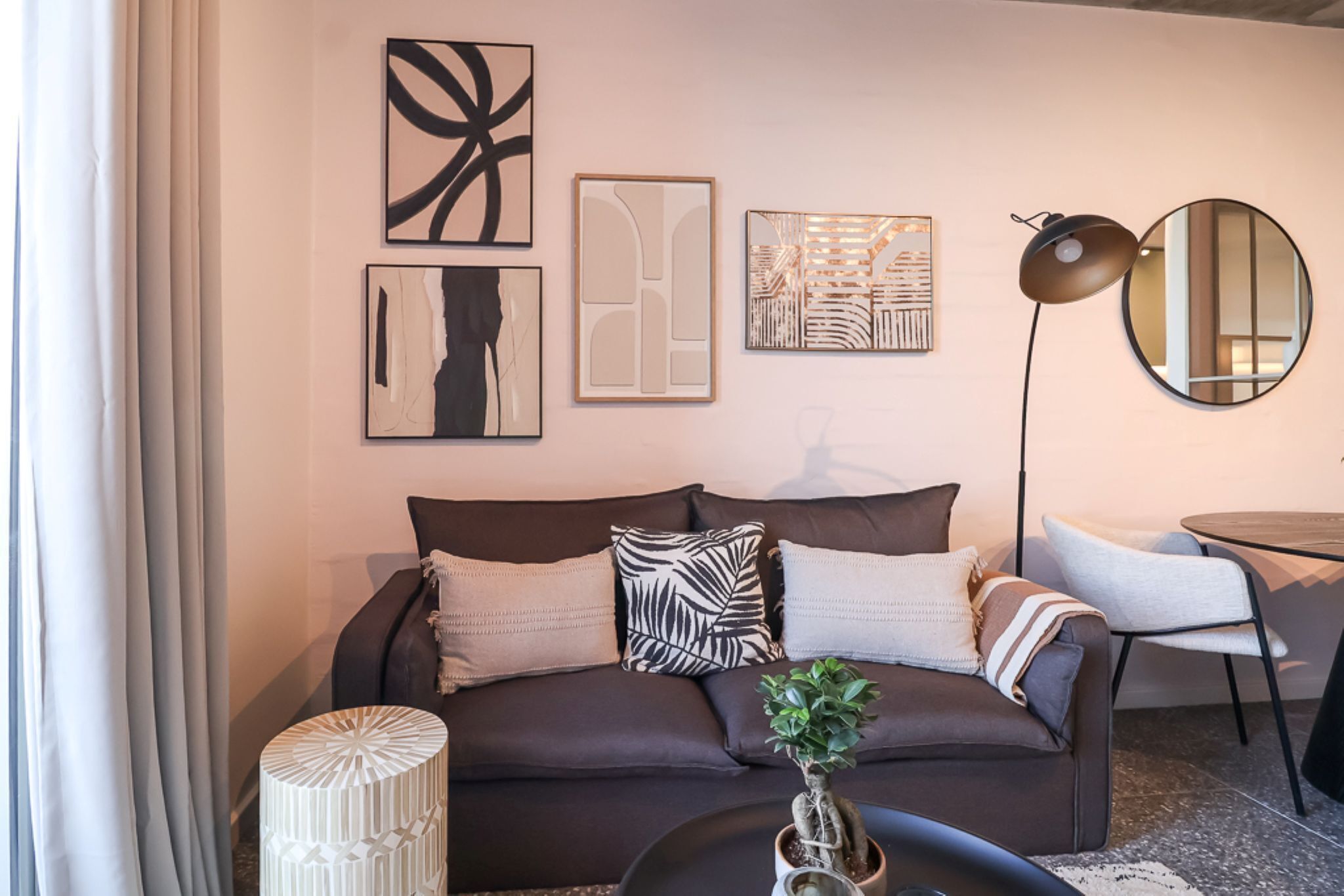 Ein modernes Wohnzimmer mit einem dunkelbraunen Sofa, einem runden Couchtisch und einer Topfpflanze. An den Wänden befinden sich abstrakte Kunstwerke, und ein Stehlampen sorgt für Licht.