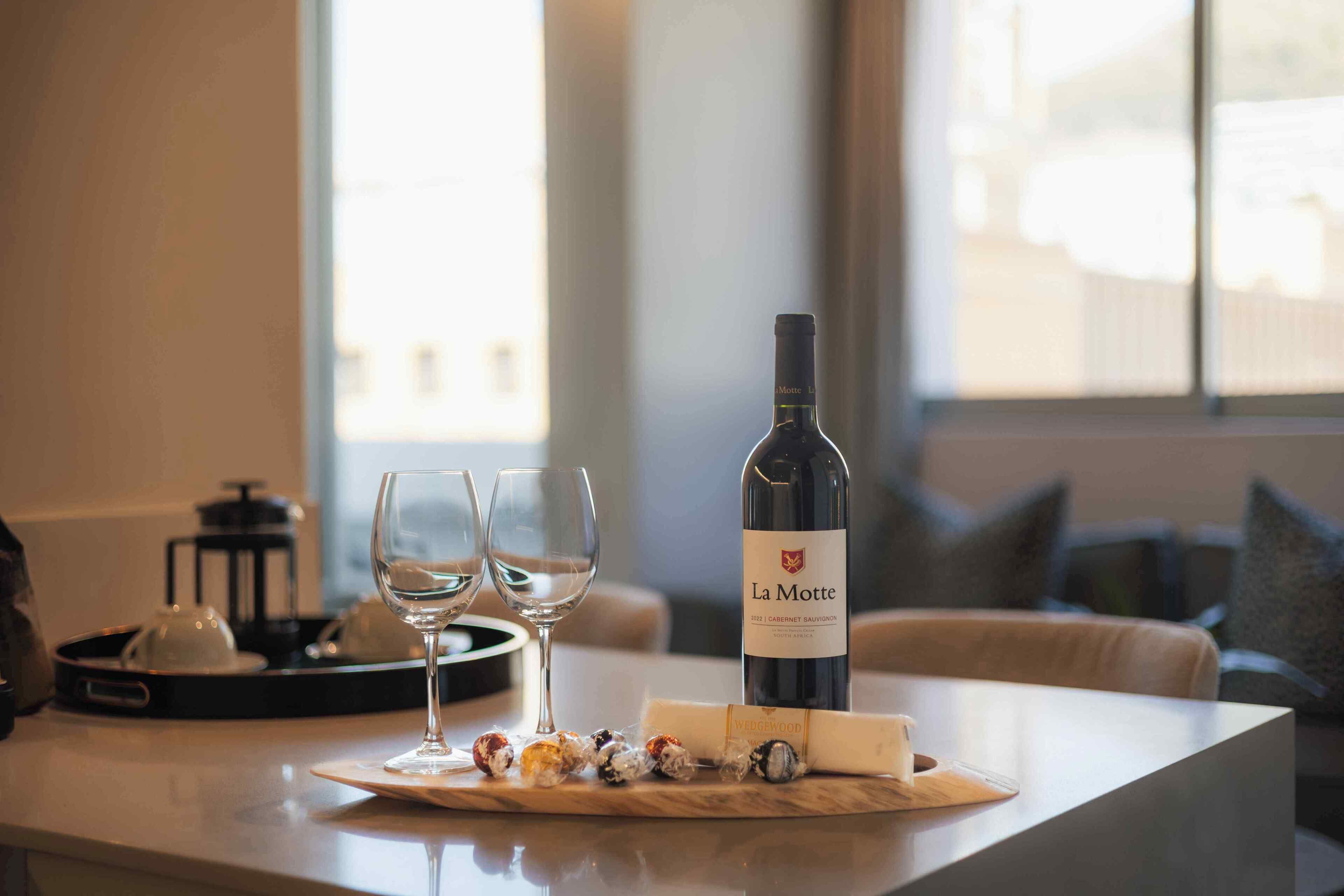 Una botella de vino La Motte, dos copas y una bandeja con bombones sobre una mesa en una habitación luminosa con vistas al exterior.