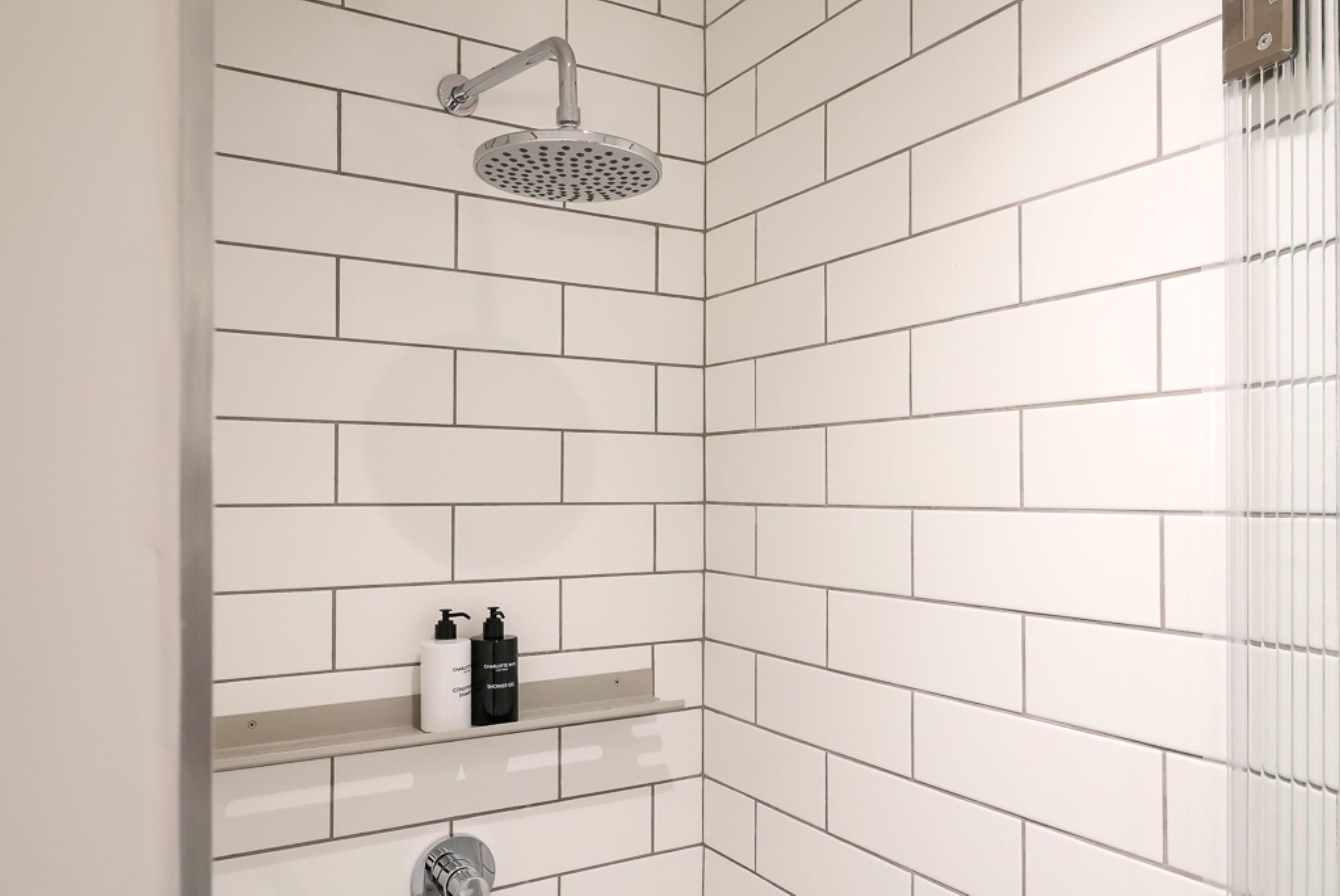 A corner shower with white tiles, a handheld showerhead, and a shelf with toiletries.
