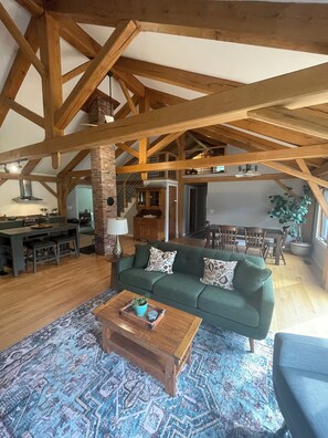 A spacious living room with a green sofa, wooden coffee table, and patterned rug. A kitchen with a brick wall and wooden beams is visible in the background. - Cozy 3-bedroom post and beam house in charming Fairfax (Fairfax)
