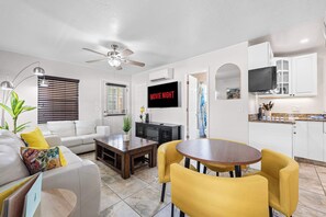 A modern living room with a sofa, coffee table, and yellow chairs. A TV is mounted on the wall, and there is a ceiling fan.