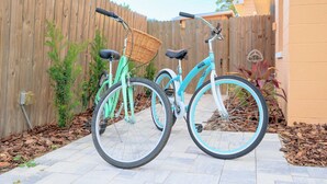 Two bicycles parked in a backyard with a wooden fence, a tiled patio, and some plants.