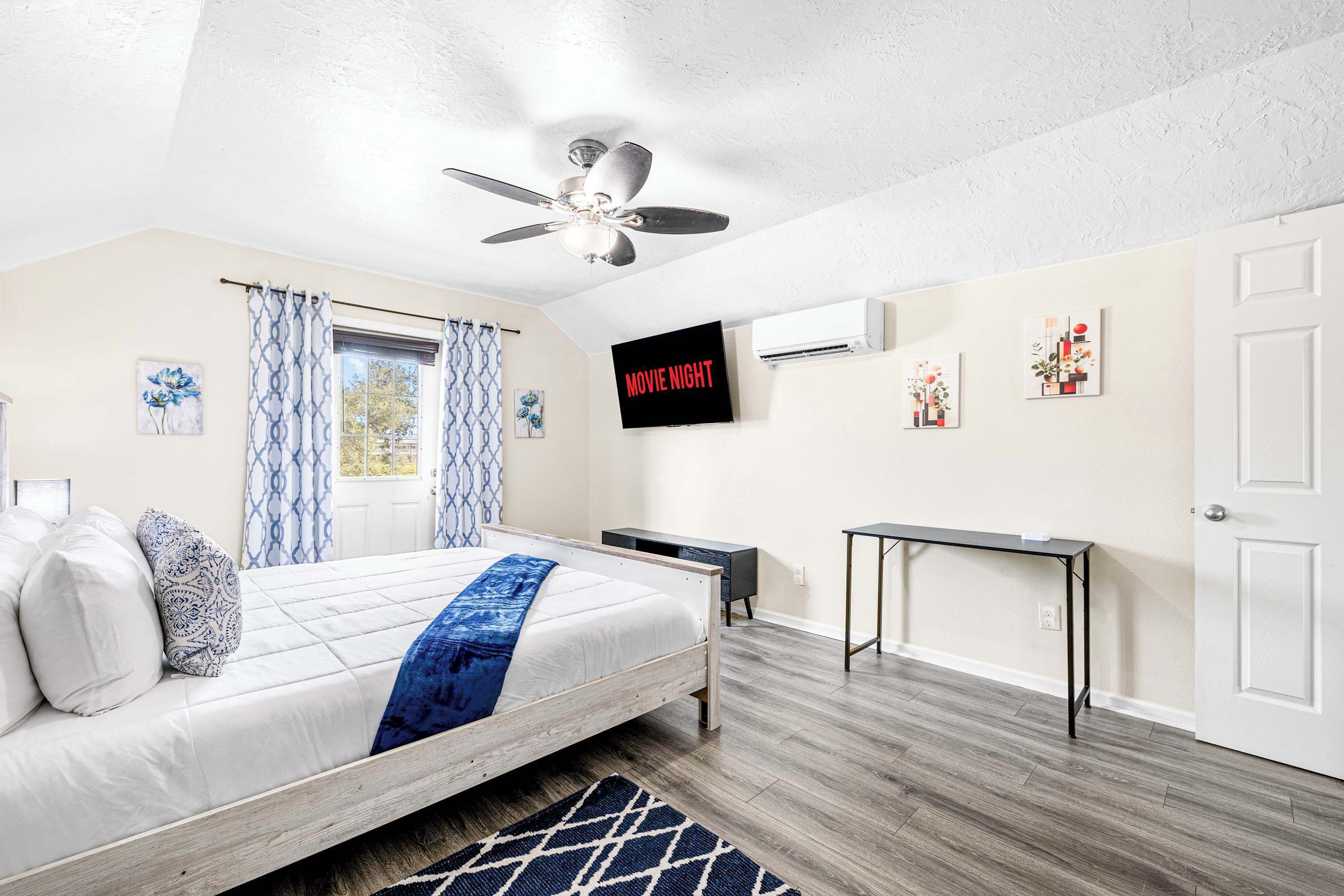 A bedroom with a bed, a ceiling fan, a TV, an air conditioner, a desk, and a door.