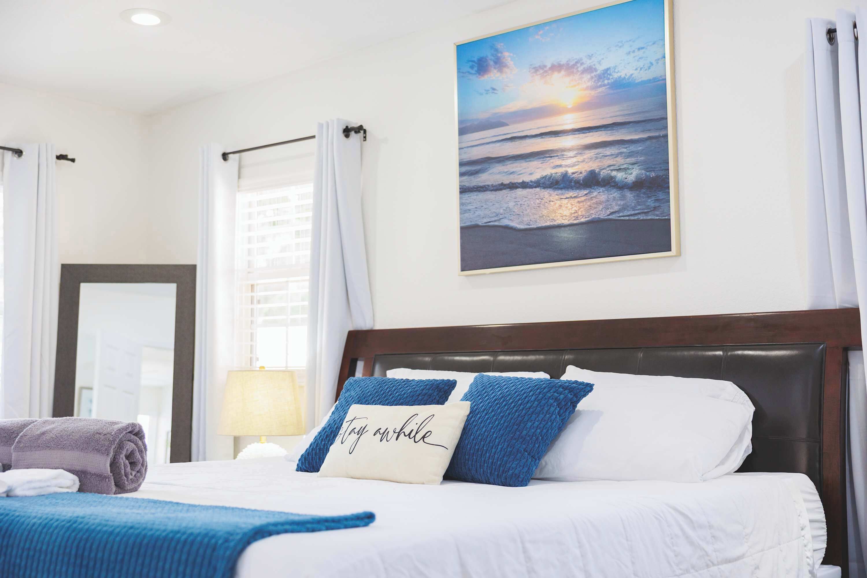 A hotel room with a bed, a painting of a sunset over the ocean, and a window with curtains.